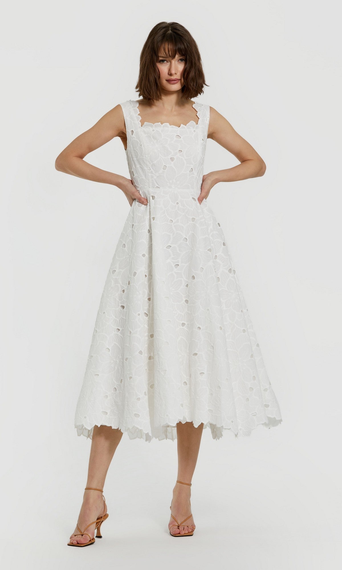 A woman stands with her hands on her hips, wearing the Mac Duggal 69096 Midi Party Dress in whiteโa sleeveless dress featuring eyelet and floral embroidery with a scalloped necklineโpaired with strappy heels against a plain background.